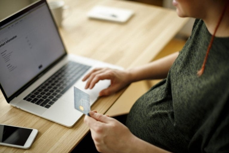 A person is sitting at a wooden table with a laptop open in front of them. They are holding a credit card in their right hand and typing on the laptop with their left hand, possibly browsing for products on Amazon. A smartphone and a cup are also on the table.