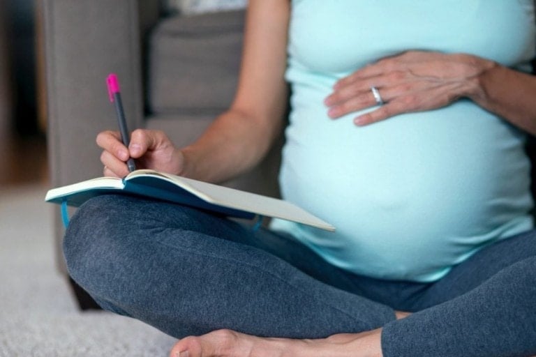 Pregnant woman writing a birth plan in a notebook while sitting cross-legged on the floor