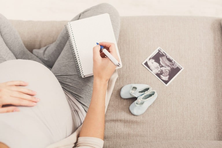 A person is sitting on a sofa with a notebook and pen in hand, jotting down notes, possibly listening to a birth plan podcast. They appear to be pregnant, with one hand resting on their belly. Next to them are a pair of baby shoes and an ultrasound image. The setting is cozy and relaxed.