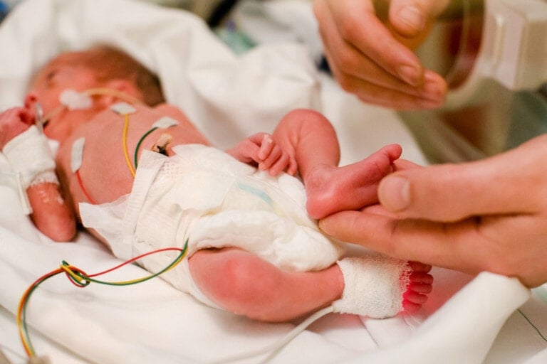 A newborn baby lies in an incubator while connected to various medical monitors. The baby is wearing a diaper, and an adult's hand is gently holding the baby's foot. The baby has monitoring pads and wires attached to its body, highlighting the critical care environment where NICU gifts can make a difference.