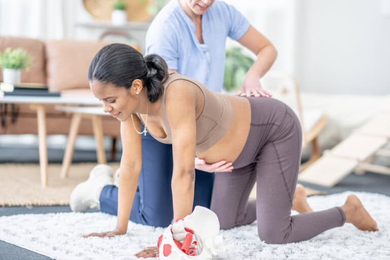 A pregnant woman performs a hands-and-knees exercise on a rug with the assistance of a physical therapist. The setting appears to be a home with a couch and potted plants in the background.