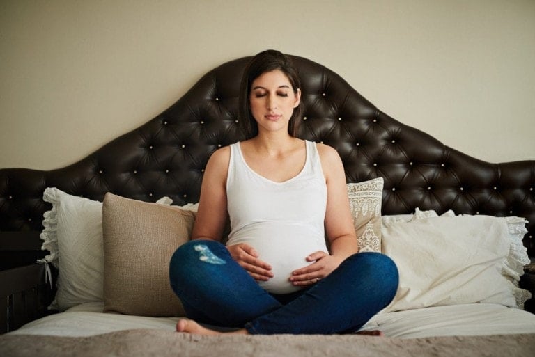 A pregnant woman sits cross-legged on a bed, eyes closed and hands resting on her belly, absorbed in a calming hypnobirthing podcast. She wears a white tank top and blue jeans. The bed has white pillows and a dark, tufted headboard. The room is softly lit.