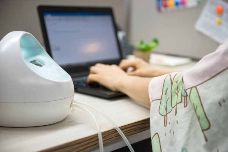 A person using a laptop at a desk while a breast pump, compliant with the Pump Act, is placed nearby. The individual appears to be working in an office setting and is wearing a cover for privacy. A green plant and various office supplies are visible in the background.