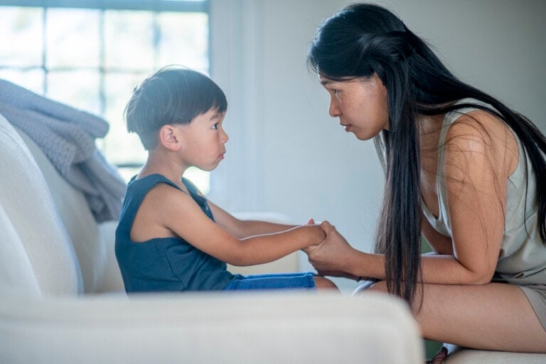 A woman with long hair kneels in front of a young boy seated on a couch. They are holding hands and looking into each other's eyes, engaged in tough conversations. The background features a window with a light curtain partially drawn aside.