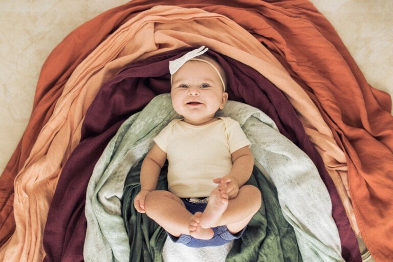 A baby, emblematic of a tender rainbow baby story, wears a white headband and light-colored onesie while lying on a layered, colorful arrangement of cloths in shades of orange, maroon, green, and beige. The baby is smiling and looking up.