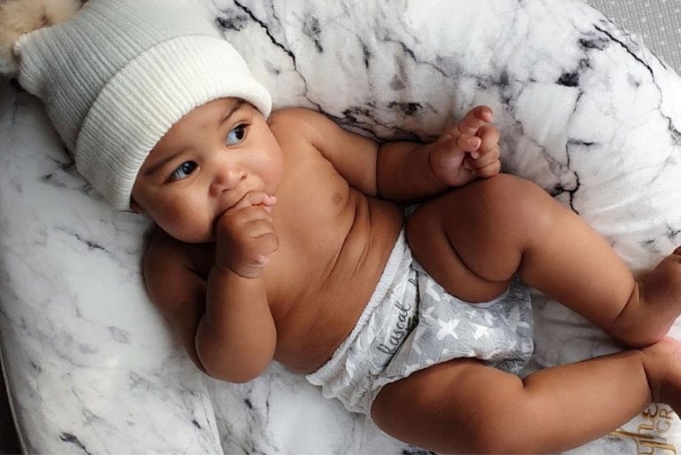 A baby is lying on a plush, white and gray marble-patterned blanket. The baby is wearing a white knit hat with a pom-pom on top and Rascal + Friends training pants with star patterns. With one hand in their mouth, the baby looks off to the side.
