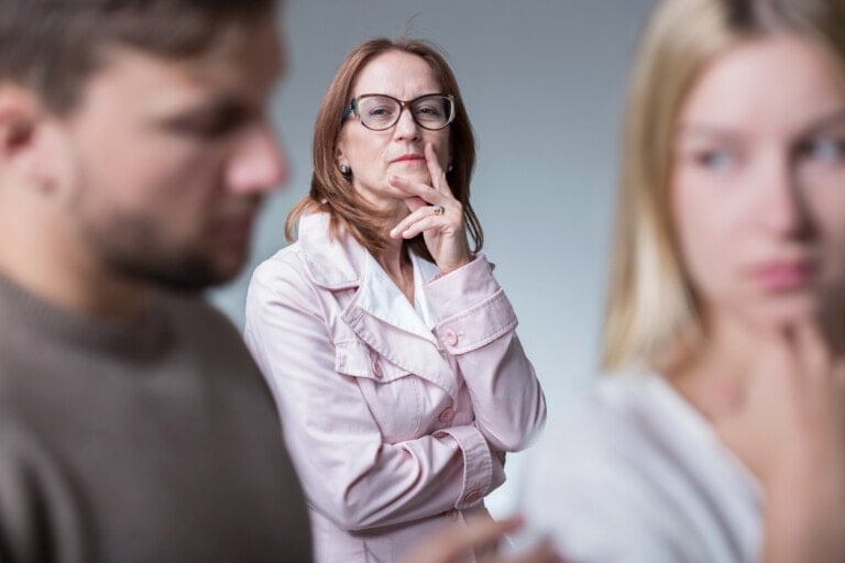 A middle-aged woman with glasses stands thoughtfully with her hand on her chin. In the foreground, a man and his MIL are slightly out of focus; the man looks downward and the mother-in-law looks sideways, both appearing concerned.