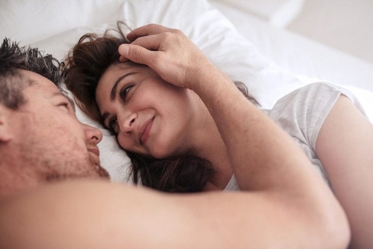 A man and woman lie in bed facing each other. The man gently touches the woman's head while she smiles at him. Both appear relaxed and comfortable, suggesting a close relationship enhanced by romantic essential oils. They are on white bedding, and the woman wears a white top.