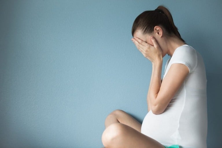 A pregnant woman sits against a blue wall with her hands covering her face, seemingly distressed. She is wearing a white t-shirt and her hair is tied back in a ponytail, highlighting the financial stress she may be experiencing during pregnancy.