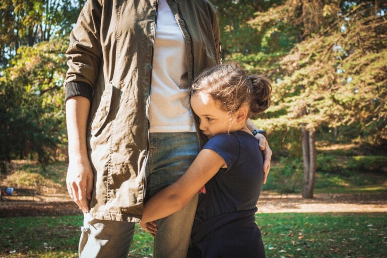 A shy kid in a navy blue shirt hugs the leg of an adult wearing a khaki jacket and jeans in a wooded park. The adult has their arm around the child, and both are standing on a dirt path with trees in the background.