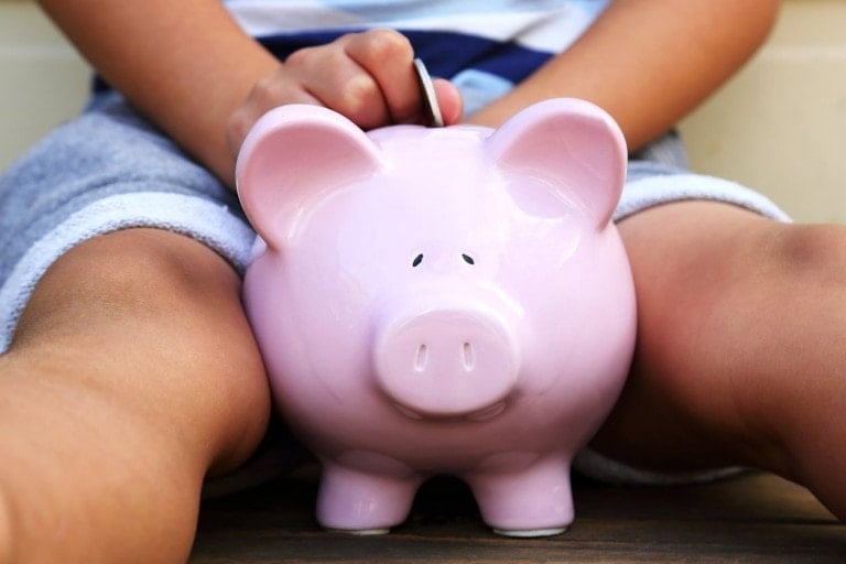 A person in grey shorts holds a pink piggy bank between their legs and is inserting a coin into it, subtly hinting at why kids should get an allowance. The background is out of focus.