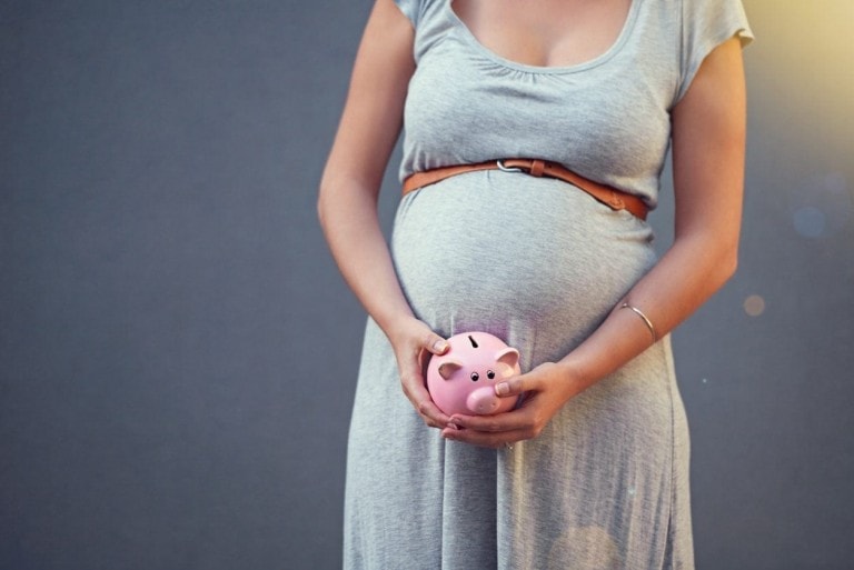 A pregnant person in a grey dress holds a small pink piggy bank with both hands in front of their belly, symbolizing financial planning for the baby. They are standing against a plain grey background, wearing a brown belt.