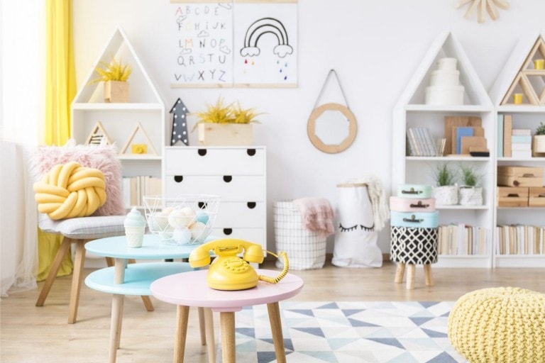 A brightly lit children's playroom with pastel-colored furniture offers perfect playroom inspiration. Two round tables, one blue and one pink, hold a yellow rotary phone and decor items. Shelving units display books and toys. A wall features a rainbow art piece and an alphabet print, sparking creativity.