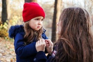 A young child wearing a red beanie and blue coat holds hands with an adult who has long hair. The child appears upset, looking at the adult in an outdoor setting with fallen leaves on the ground, as if they're trying to get toddlers to listen without resorting to bribes and threats.