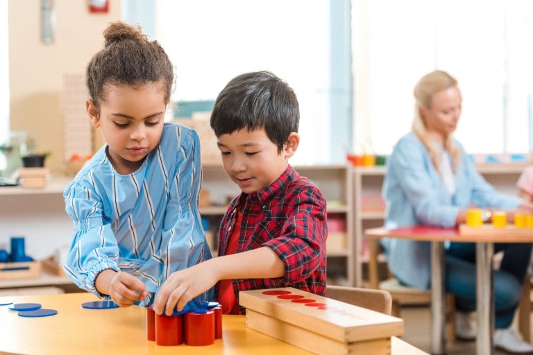 Two young children are playing together at a table in a classroom, demonstrating social skills for kids. One child places blue disks into a red container while the other watches. In the background, a teacher and another child are engaged in an activity, providing more social skills examples.