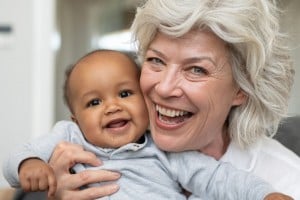 An older woman with gray hair, possibly known by sweet grandma names, is holding a smiling baby. Both have wide smiles and are looking directly at the camera. The woman is wearing a white top, and the baby is dressed in a gray sweater. They appear to be indoors.
