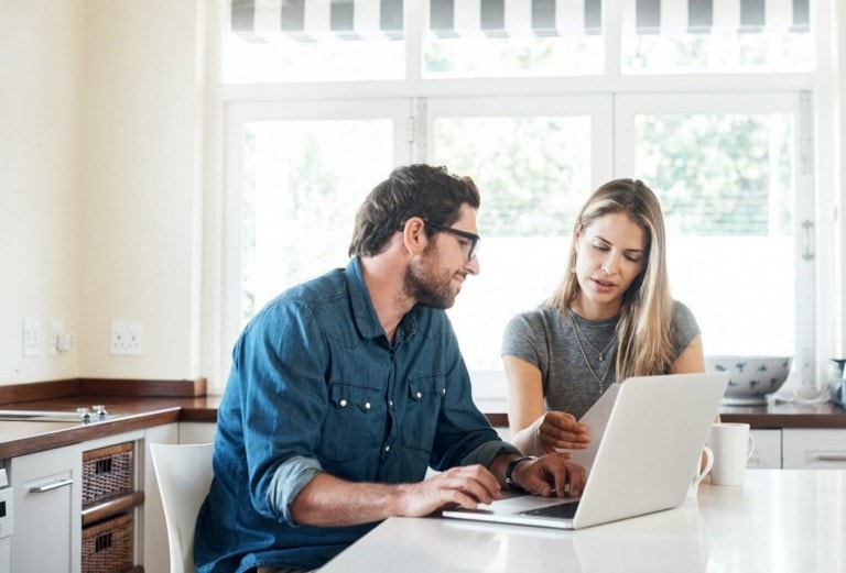 A man and woman sit at a kitchen table, both looking at a laptop screen. The man, wearing glasses and a blue denim shirt, is typing on the laptop. The woman, in a gray shirt, holds a document and appears engaged in the discussion about their money date.