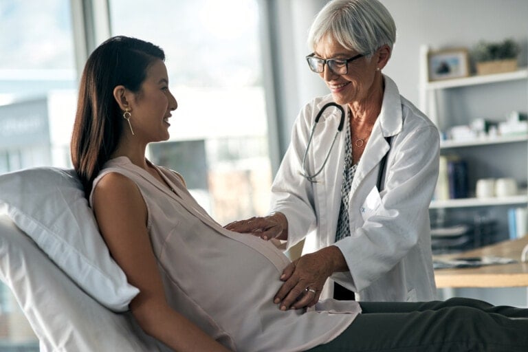 A pregnant woman in a sleeveless blouse is sitting on a medical examination table, smiling at an older female doctor with a stethoscope who is touching her belly. They engage in shared decision making with your doctor, surrounded by the bright light of the medical office filled with windows and equipment.