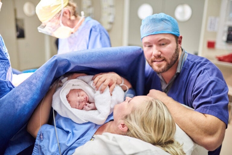 A mother, lying in a hospital bed, holds her newborn baby wrapped in a white blanket. A man in blue scrubs and a surgical cap, possibly the father, stands beside her. Medical personnel are present in the background of the delivery room, where emotions run high despite potential PTSD lingering for some families.