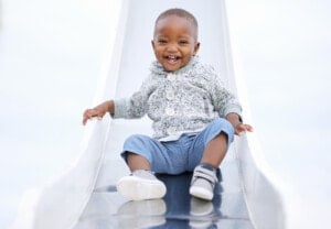A small child named Zane, wearing a gray sweater, blue pants, and white shoes, is sitting on a slide. The child is smiling and holding onto the sides of the slide with their hands.