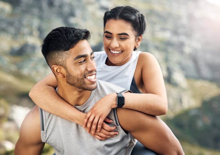 A couple outdoors, with a woman playfully getting a piggyback ride from a man. They are both smiling in an open, mountainous setting. The woman is wearing a smartwatch.