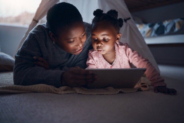An adult and a child are lying on the floor inside a cozy room, leaning on a yellow blanket and looking at a tablet together. The adult is wearing a gray sweater, and the child is wearing a pink top with hair styled in multiple ponytails as they explore popular bedtime stories.