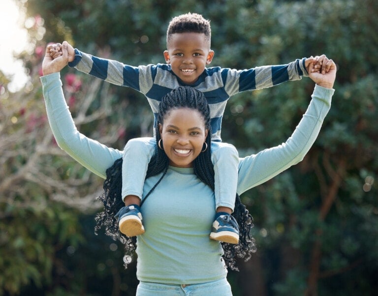 A kind-hearted boy in a striped shirt smiles as he sits on a woman’s shoulders. Both have their arms raised and are holding hands, radiating joy. The woman, dressed in a light blue long-sleeve top, stands amidst vibrant trees and foliage that provide a beautiful backdrop to their happiness.