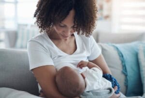 A woman with curly hair is sitting on a couch, breastfeeding a baby wrapped in a blanket treated with flame retardants. She is looking down at the baby, and the background shows a softly lit room with cushions.