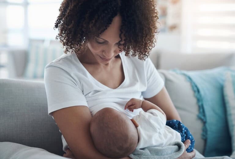 A woman with curly hair is sitting on a couch, breastfeeding a baby wrapped in a blanket treated with flame retardants. She is looking down at the baby, and the background shows a softly lit room with cushions.