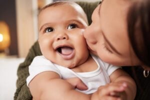 A close-up image of a smiling baby wearing a white shirt being kissed on the cheek by an adult. The baby looks joyful with mouth open and wide eyes. The background is softly blurred, focusing attention on the baby, who might just grow up to have one of those charming nicknames for boys, and the loving gesture.