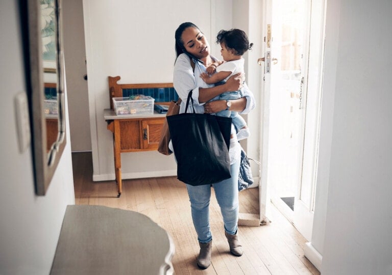 A woman stands in an entryway holding a child in one arm and a large black breast pump bag in the other. She is dressed casually in jeans and a light-colored shirt. The room is bright, with a small table against the wall and a wooden chair nearby. The front door is open.