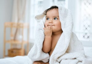 A 9-month-old baby sits on a bed, wrapped in a white towel with the hood covering its head. The baby has a curious expression, with one hand covering its mouth. The setting appears to be a bright and airy room with blurred background decor, suggesting it's time for the 9-month-old's bedtime routine.