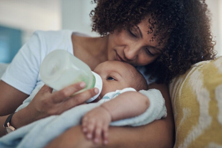 A woman with curly hair is holding and bottle-feeding a baby wrapped in a light blue blanket. She is sitting on a couch with yellow and white patterned cushions. Despite her nursing aversion, the baby looks up at her lovingly while drinking from the bottle.