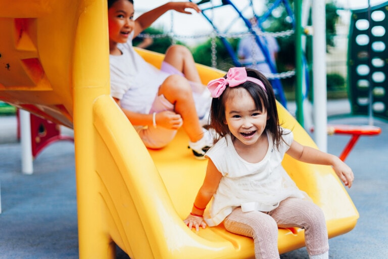 Two children are playing on a yellow slide at a playground, enjoying free summer activities for kids. The child in the foreground is a young girl wearing a pink bow in her hair and a white outfit, smiling at the camera. The child in the background, also smiling, is sliding down behind her.
