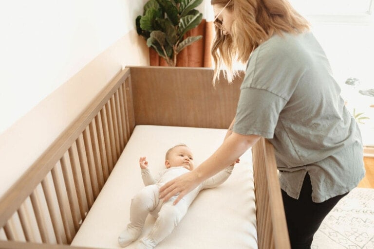 A woman places a sleeping baby dressed in white on their back in a wooden crib. The room has a large plant in the corner and light coming through a window, creating a bright and airy atmosphere, ensuring the baby is safe and serene.