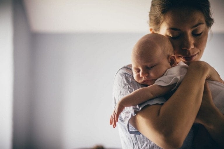 A woman holds a sleeping baby close to her chest in a softly lit room with light-colored walls. Her eyes are closed as she gently cradles the infant, who is dressed in a white outfit. This serene moment embodies the essence of stories shared on the "Podcast MotherhoodTies.