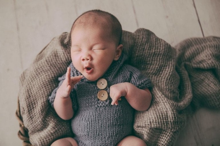 A baby wearing a gray knit onesie with two buttons on the front is lying in a soft, brown blanket. The baby has their eyes closed and mouth slightly open, with one hand raised. As you imagine naming this little one, consider some of the coolest baby boy names for an extra special touch.