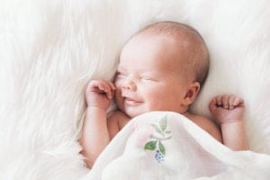A smiling baby is lying on a fluffy white surface, covered partially by a light blanket with a floral design. The baby, often called by sweet nicknames, has eyes closed and both hands near the face.