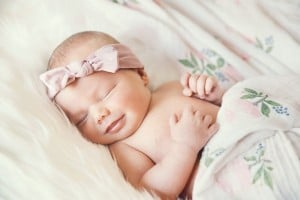 A sleeping baby with a pink bow headband is lying on a soft, white surface. Partially covered by a floral-patterned blanket, this serene infant resembles the kind of cool baby girl names that exude both elegance and tranquility.