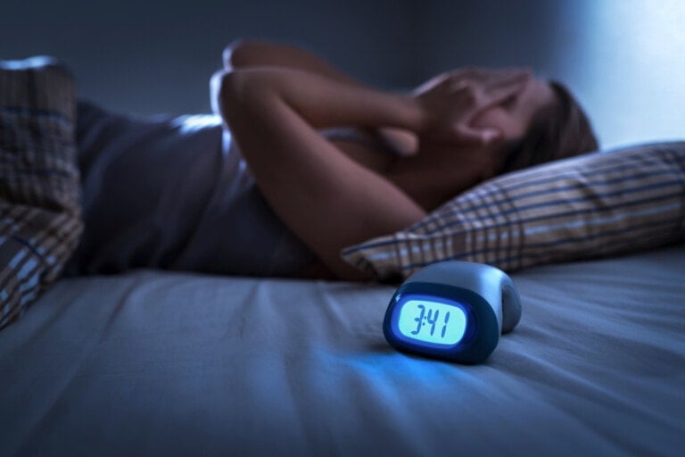 A person lies in bed with hands covering their face, appearing to experience postpartum insomnia. An alarm clock on the bedside table shows the time as 3:41, emitting a blue light in a dark room.