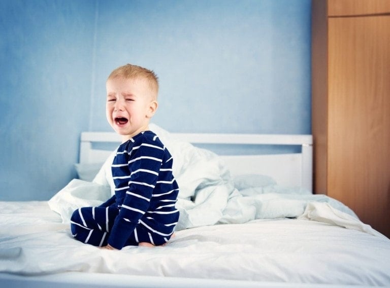 A young child wearing blue and white striped pajamas sits crying on a bed, possibly due to bedwetting. The child is positioned towards the left side of the white bed, with crumpled bedding visible behind them. The room has light blue walls and a wooden wardrobe in the background.