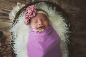 A sleeping baby wearing a purple swaddle and a pink flower headband rests in a fluffy-lined basket. The baby, who might have one of those charming girl names that start with Q, appears content and peaceful in this cozy setting.