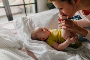 A person and a baby are on a bed. The person is kissing the baby's foot while holding their leg. The baby, possibly still figuring out its sleep schedule, is lying on its back wearing a yellow onesie and appears to be smiling. The bed has a striped sheet, and there are large windows in the background.