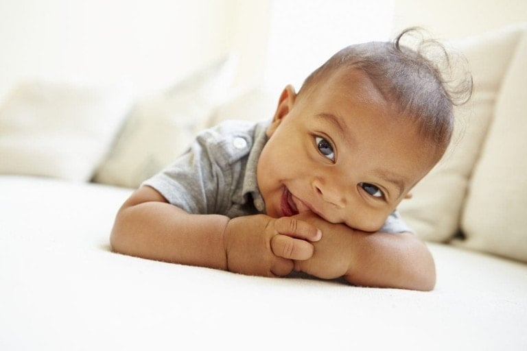 A baby is lying on their stomach, resting their head on crossed arms. The baby is wearing a grey shirt and smiling toward the camera, enjoying the benefits of tummy time. The background consists of light-colored, out-of-focus cushions.