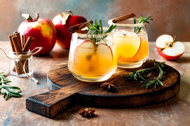 Two glasses of apple cider cocktails with ice, garnished with apple slices, rosemary sprigs, and cinnamon sticks on a wooden serving board. Fresh apples, a bundle of cinnamon sticks, and star anise are placed around the board—perfect for your fall mocktail recipes collection.