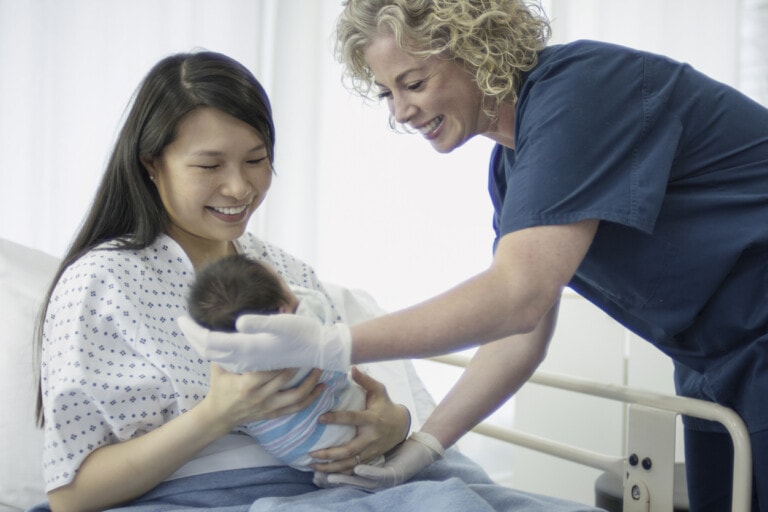 A woman sitting in a hospital bed holds a newborn baby wrapped in a blanket. A postpartum nurse, standing beside the bed, is smiling and adjusting the baby. The woman and the nurse are both smiling.