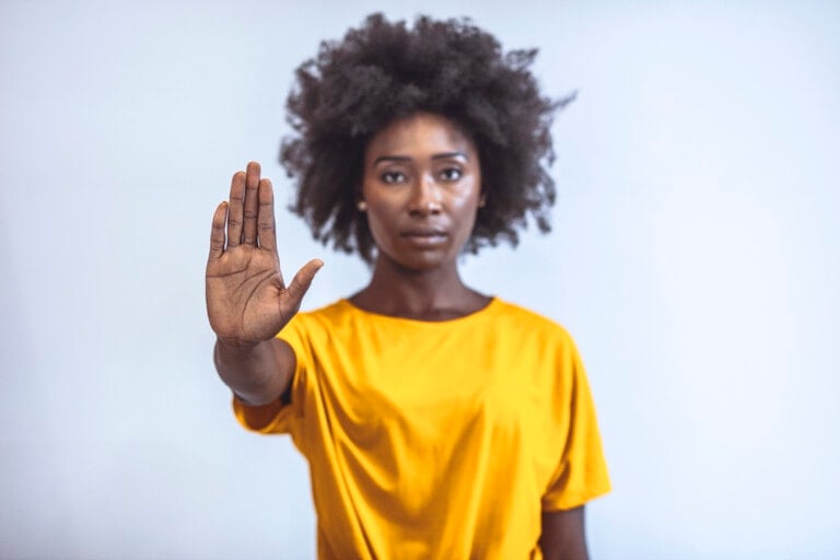 A person with an afro hairstyle stands against a plain background, wearing a bright yellow t-shirt. They have their right arm raised with their palm facing outward, as if signaling to stop or halt. Their expression seems to say, "Stop asking women.