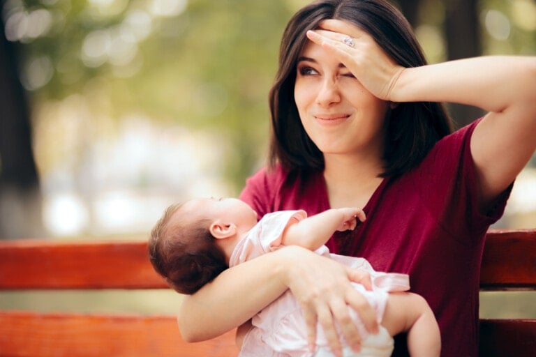A woman wearing a maroon shirt holds a baby in her arms while sitting on a wooden bench outdoors. The woman appears amused, possibly due to mommy brain, and is wiping her forehead with her hand. The background is blurred with greenery.