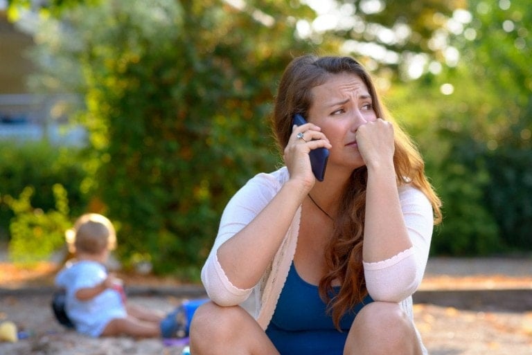 A woman with long hair is sitting outdoors, holding a phone to her ear with a concerned expression on her face, possibly dealing with adultery. She is wearing a light cardigan over a blue top. In the background, a child is playing, but the focus remains on the woman.