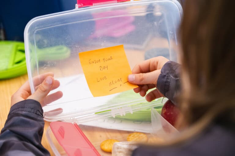 A person is holding an open lunchbox with a note inside that reads, Enjoy your day. Love, Mom. The lunchbox contains an apple, some crackers, and a sandwich. With a dark-colored sleeve visible as they lift the lid, its clear these sweet lunchbox notes for kids make school days extra special.